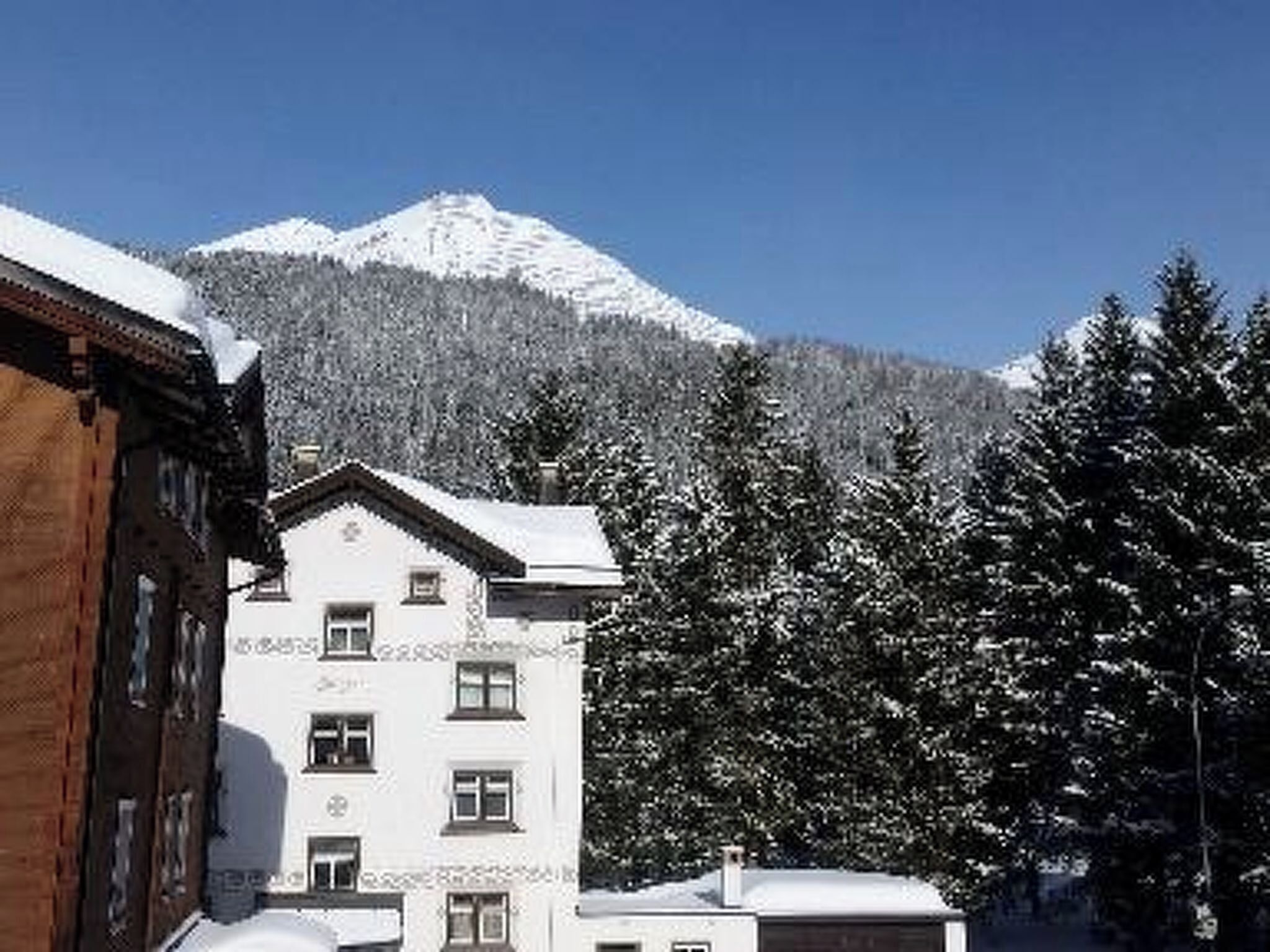 A multi-story building with a snow-covered roof, surrounded by trees and a mountain in the background.