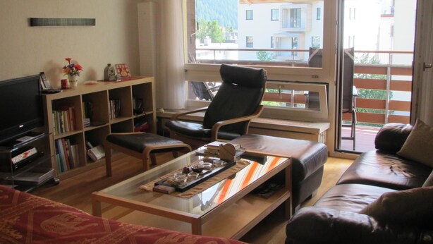 Ein Wohnzimmer mit Sofa, Sessel, Couchtisch, Fernseher, Bücherregal und einer Schiebetür zu einem Außenbereich.