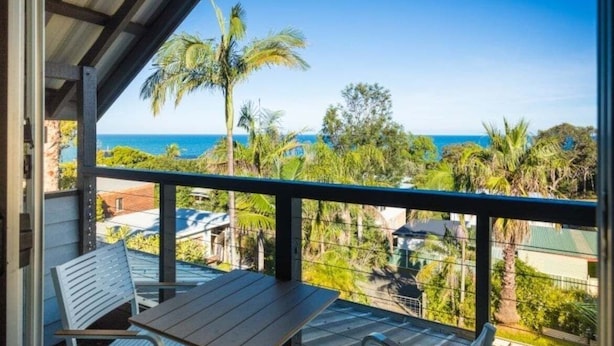Un balcon avec vue sur l’océan, des palmiers et une table en bois.