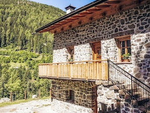 A stone building with a wooden balcony, a stone staircase, and a view of a forested hillside.