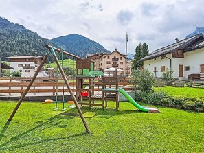 Ein Spielplatz mit Schaukeln und einer Rutsche, umgeben von Gebäuden und einem Berg im Hintergrund.