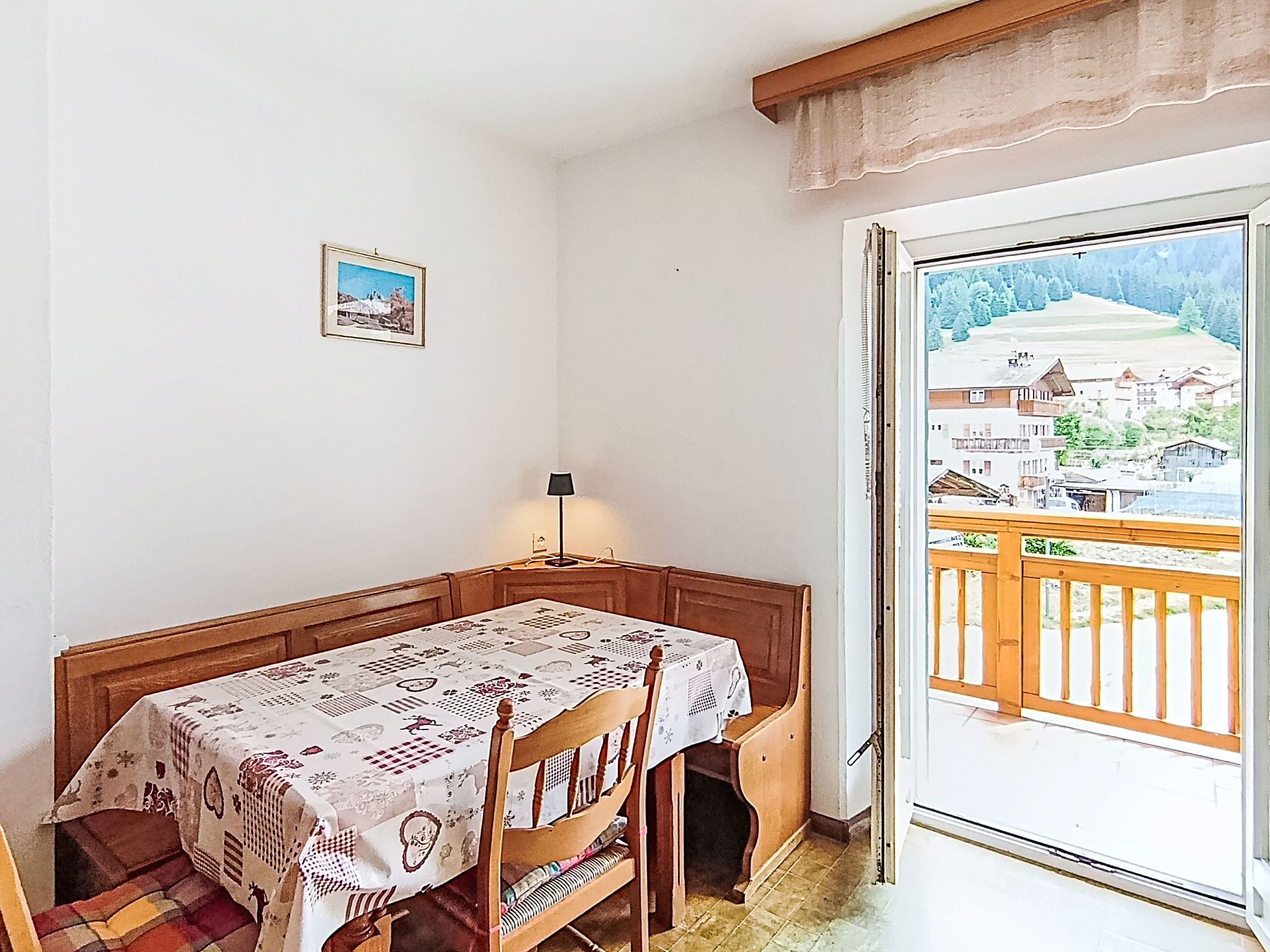 Een kamer met een houten tafel en stoelen, een lamp, een lijst met een afbeelding aan de muur, en een balkon met uitzicht op gebouwen en bomen.