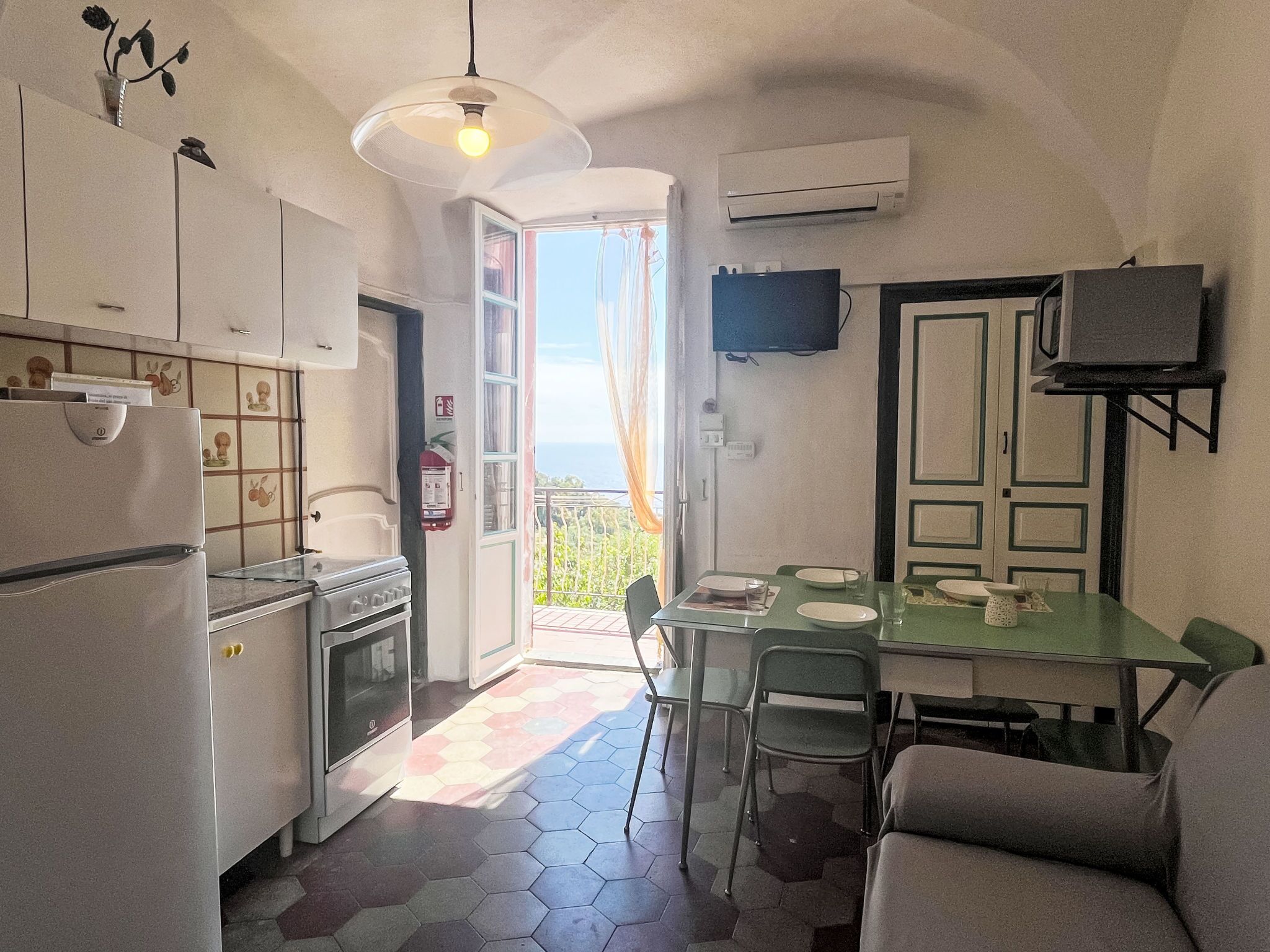 Une cuisine compacte avec un coin repas, un téléviseur fixé au mur et une vue sur l’extérieur par la porte ouverte.