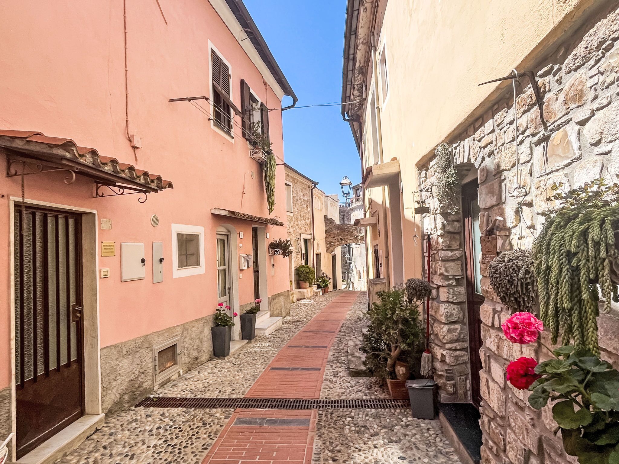 A narrow street with buildings on either side, a stone wall on the right, and a metal gate on the left.