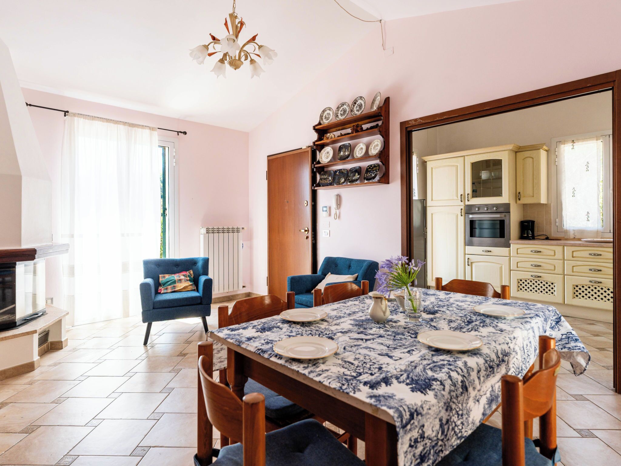 A dining area with a table set for four, a blue armchair, a sofa, and a kitchen visible through an open door.