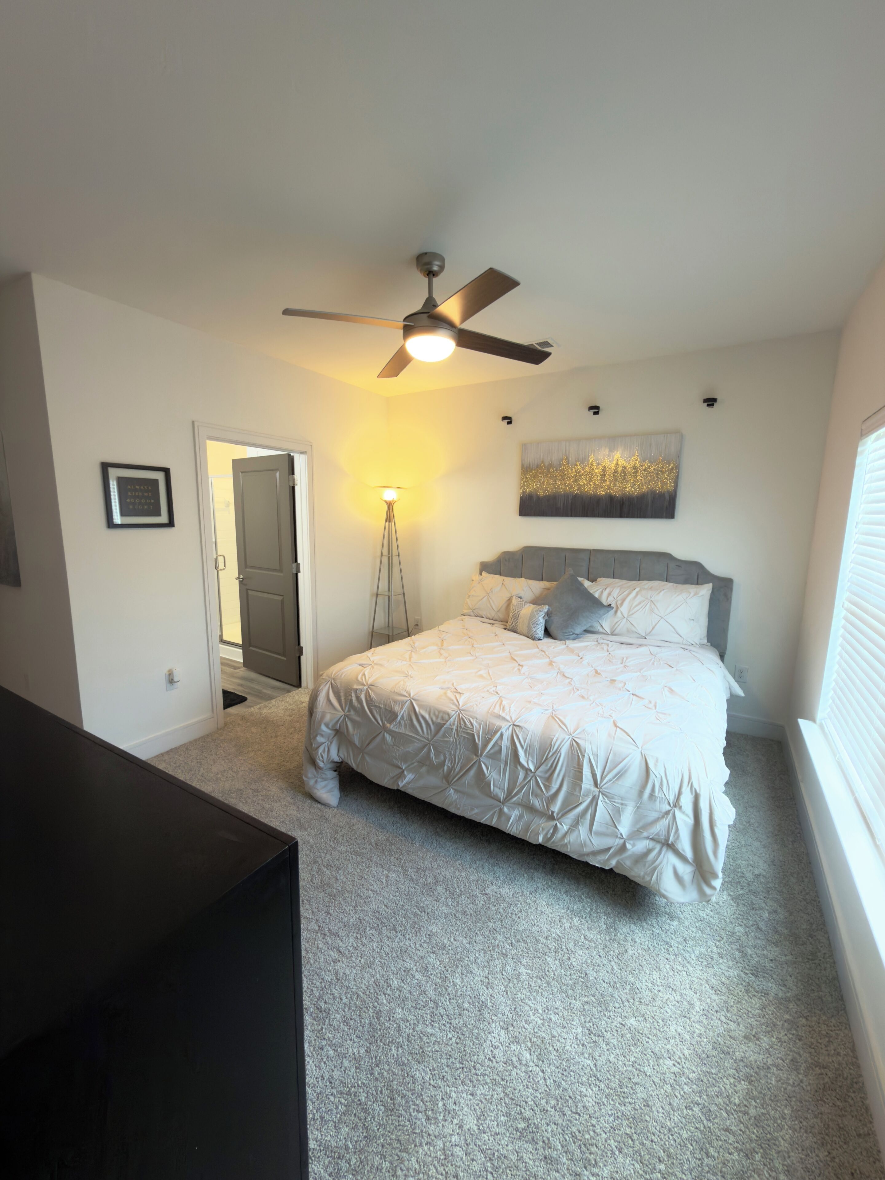 A bedroom with a bed, a ceiling fan, a floor lamp, a door, and a wall-mounted artwork.