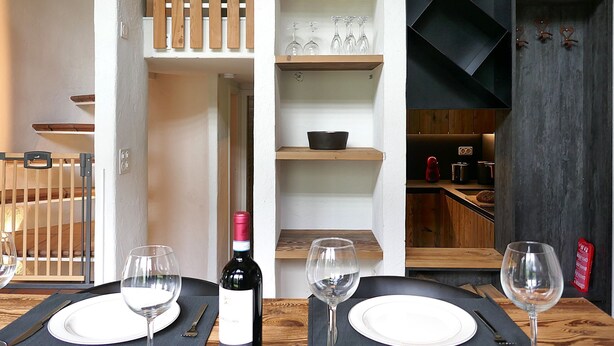 Un comedor con una mesa lista para dos personas, una botella de vino y una estantería con copas y un cuenco.