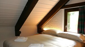 Un dormitorio con techo inclinado, una cama con ropa blanca y una ventana con cortina escocesa.