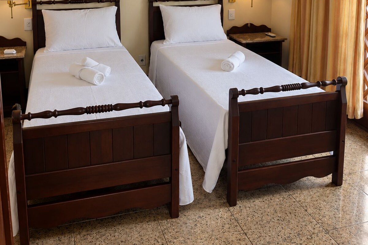 A hotel room with two wooden beds, white linens, a painting of flowers on the wall, an air conditioning unit, and a window with curtains.