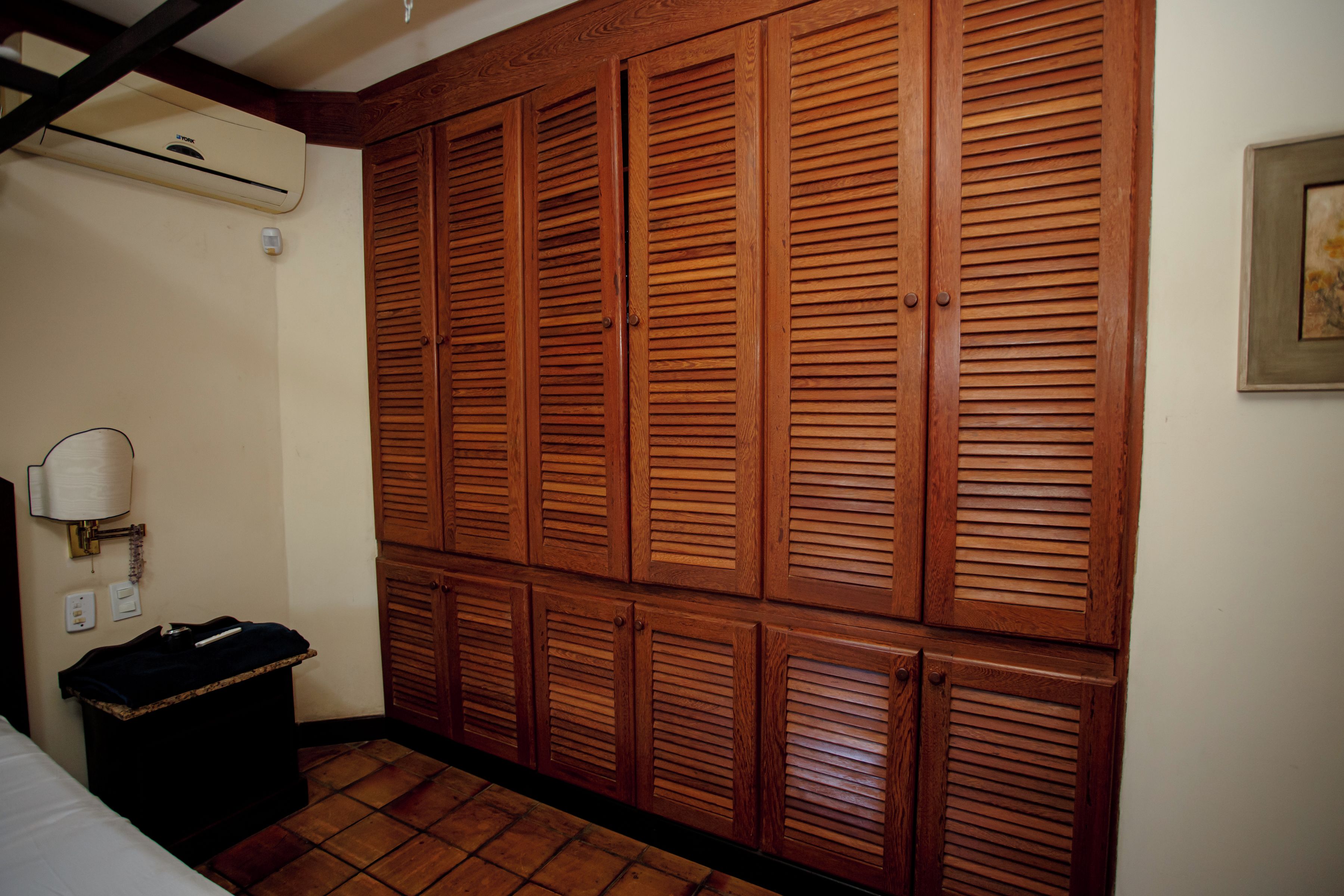 A hotel room with a large wooden wardrobe, a bedside table with a lamp, and a tiled floor.