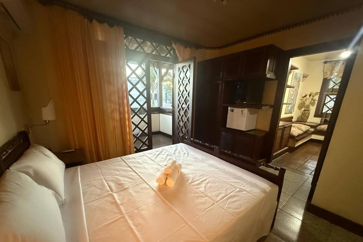 Ein Hotelzimmer mit Bett, Holz-Kopfteil, Nachttisch, Lampe, Schrank und Blick durch ein Fenster mit Vorhängen.