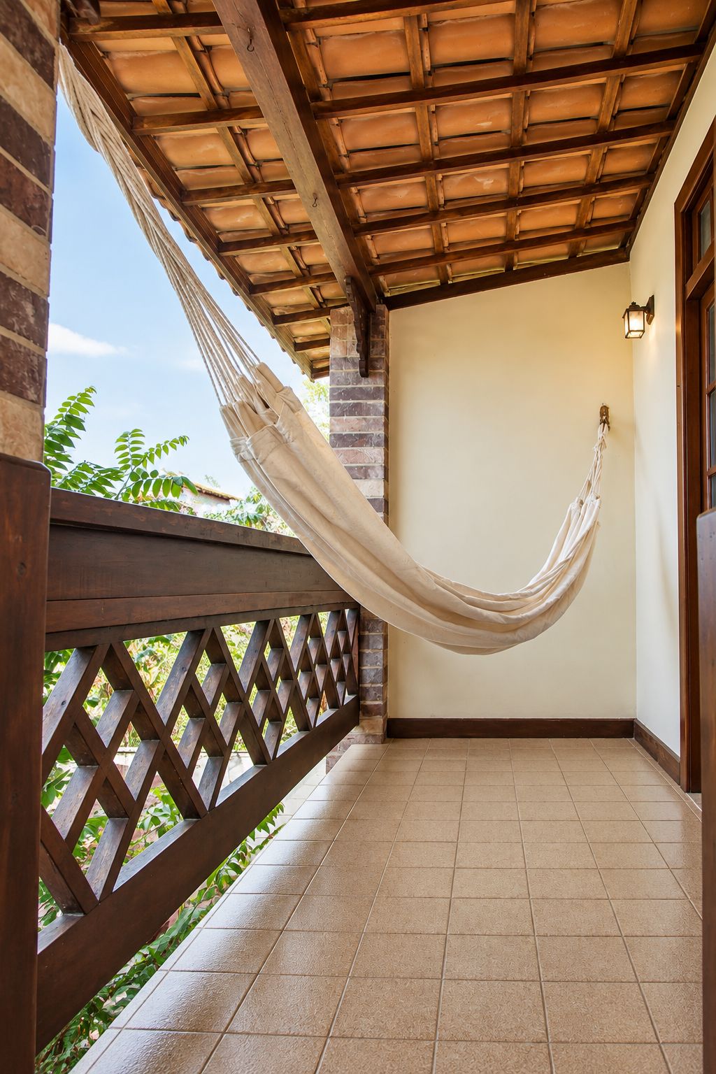 Eine Hängematte, die zwischen Holzbalken an einem Balkon gespannt ist, mit Fliesenboden und Gittergeländer.