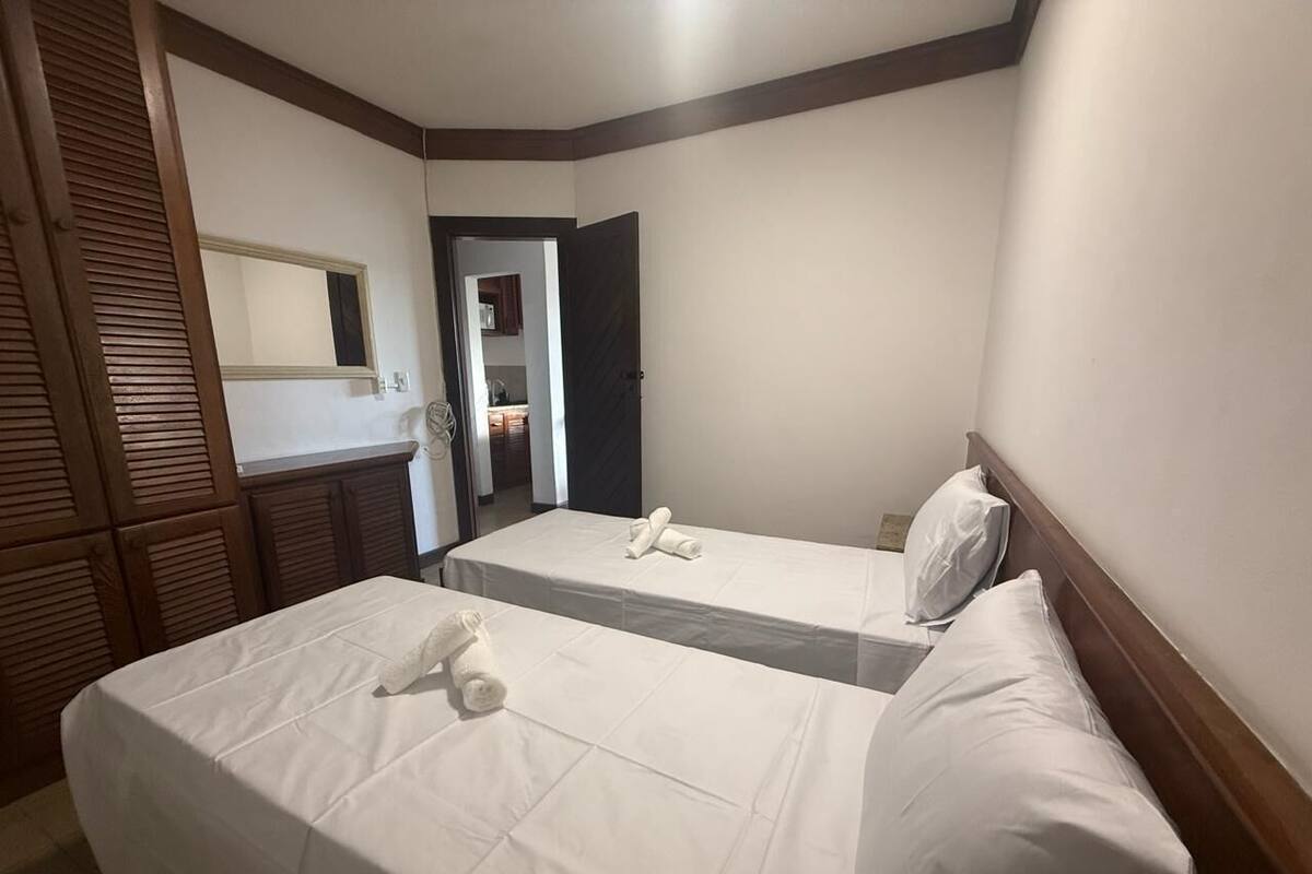 Ein Hotelzimmer mit zwei Betten, einem hölzernen Kopfteil, einem kleinen Badezimmer und einer sichtbaren Tür zu einem anderen Raum.