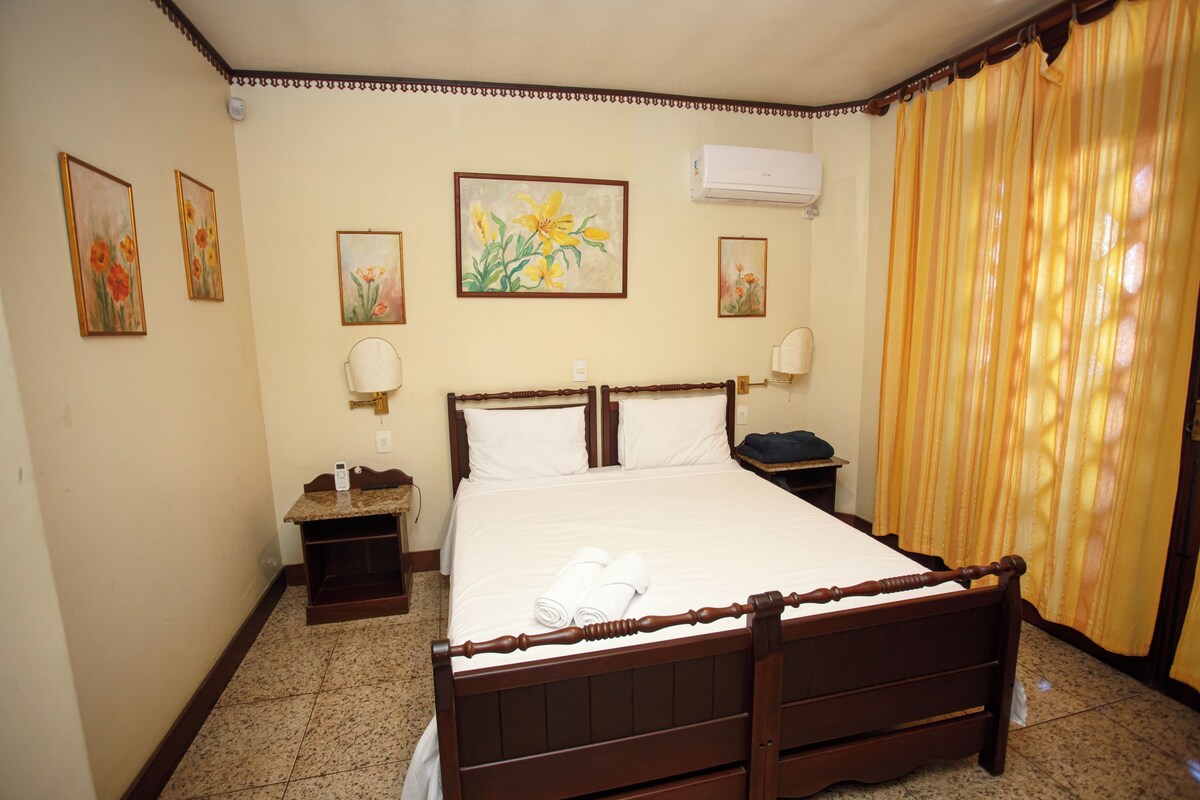 A hotel room with a wooden bed, a nightstand, a bedside table, a wall-mounted air conditioner, and a window with curtains.