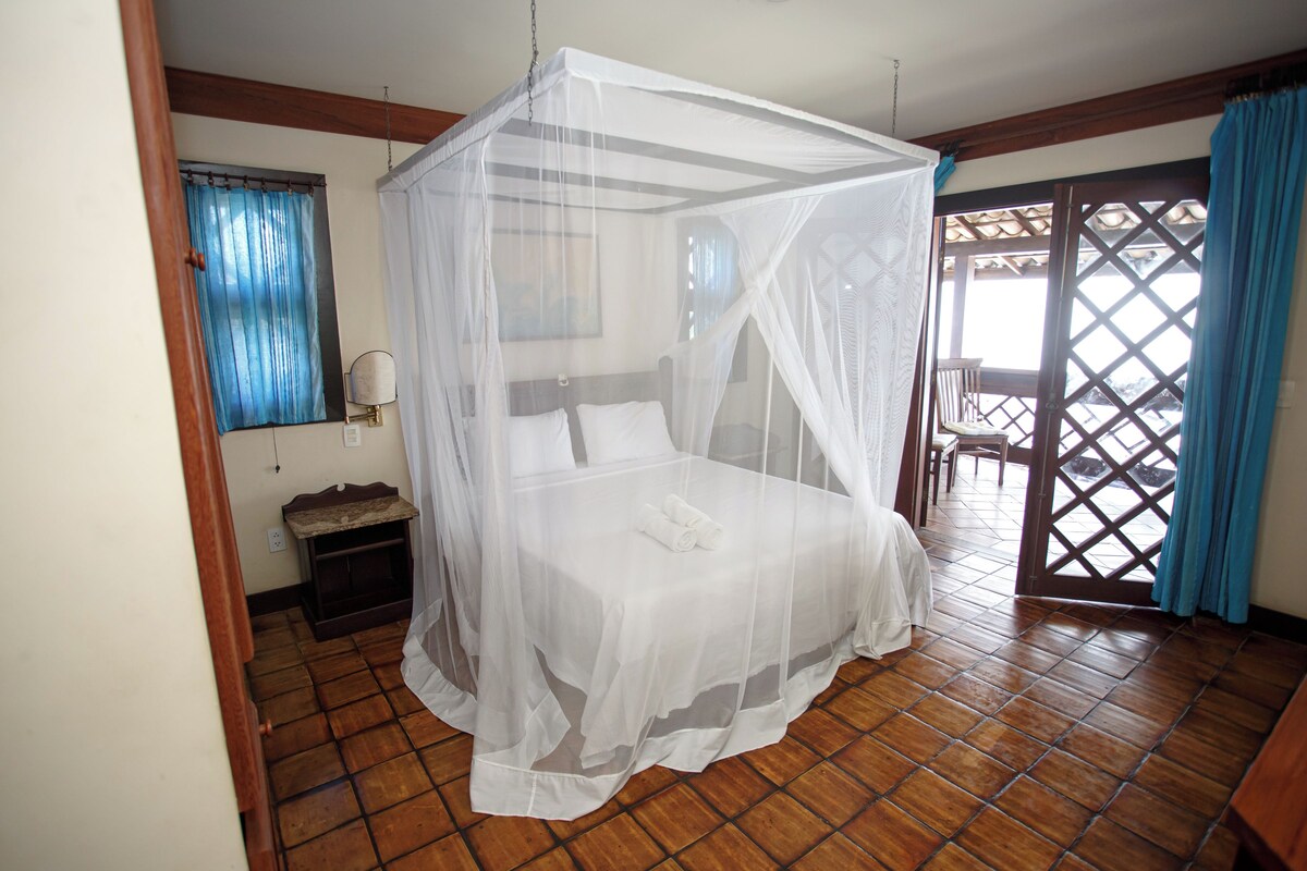 A room with a bed under a canopy, a small bedside table, a window with curtains, and a view of the outside.