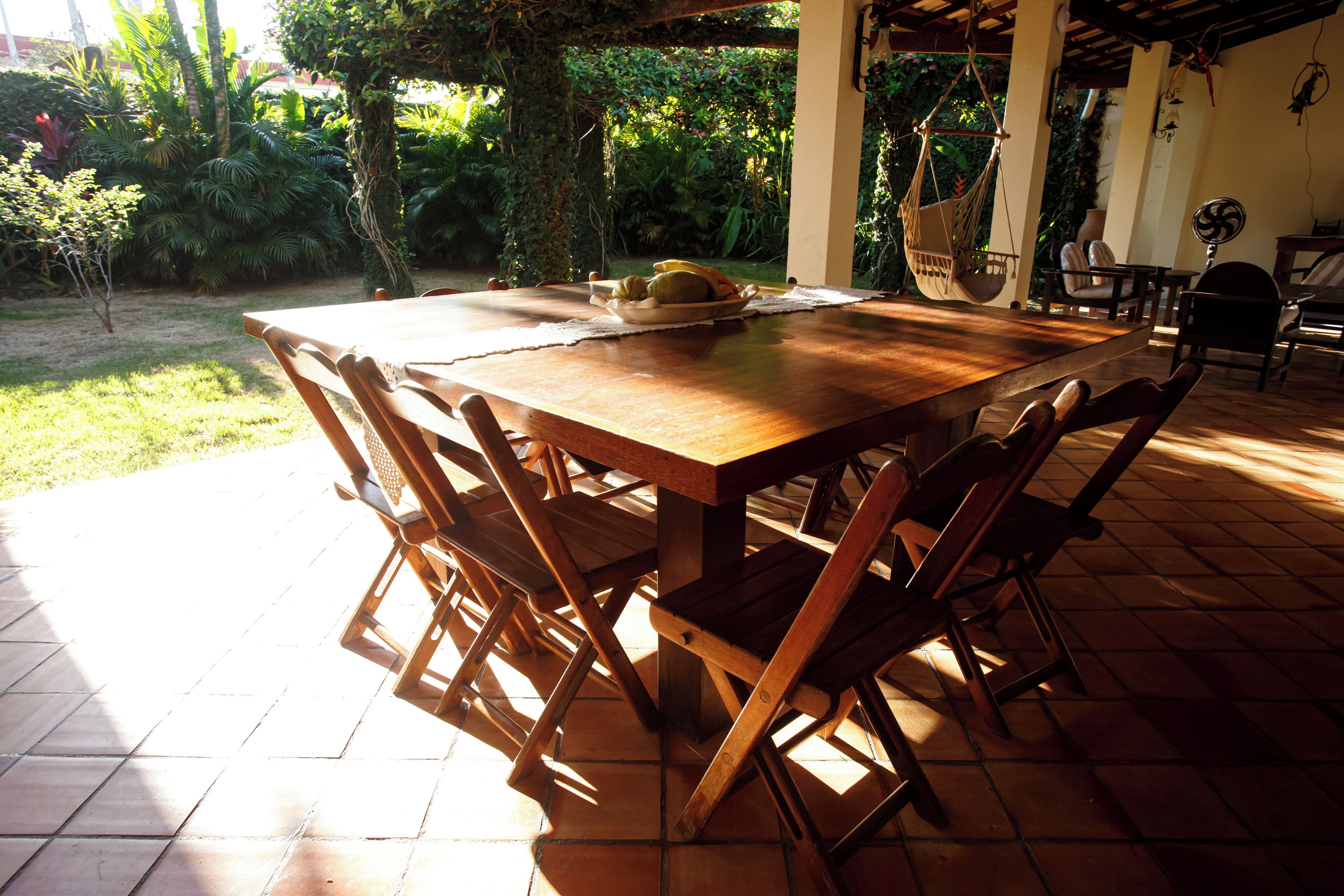 An outdoor wooden table with chairs, surrounded by a garden with various plants and a tiled floor.