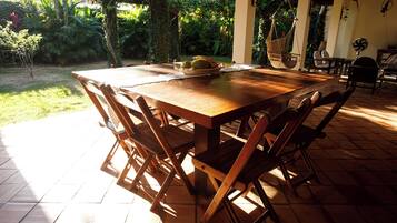 An outdoor wooden table with chairs, surrounded by a garden with various plants and a tiled floor.