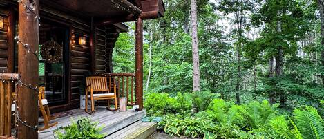 Une cabane en rondins dotée d’une véranda, d’une chaise en bois et d’un chemin bordé de pierres, le tout entouré d’une végétation luxuriante.