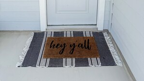 A white door with a wreath, a doormat with 'hey y'all' text, and a security camera.