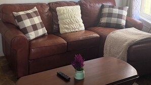 A brown leather sofa with checkered and fluffy cushions, a wooden coffee table with a remote control and a vase of purple flowers, and a painting of a beach scene on the wall.