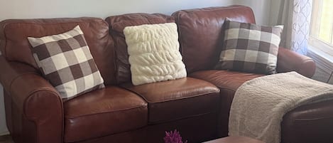 A brown leather sofa with checkered and fluffy cushions, a wooden coffee table with a remote control and a vase of purple flowers, and a painting of a beach scene on the wall.