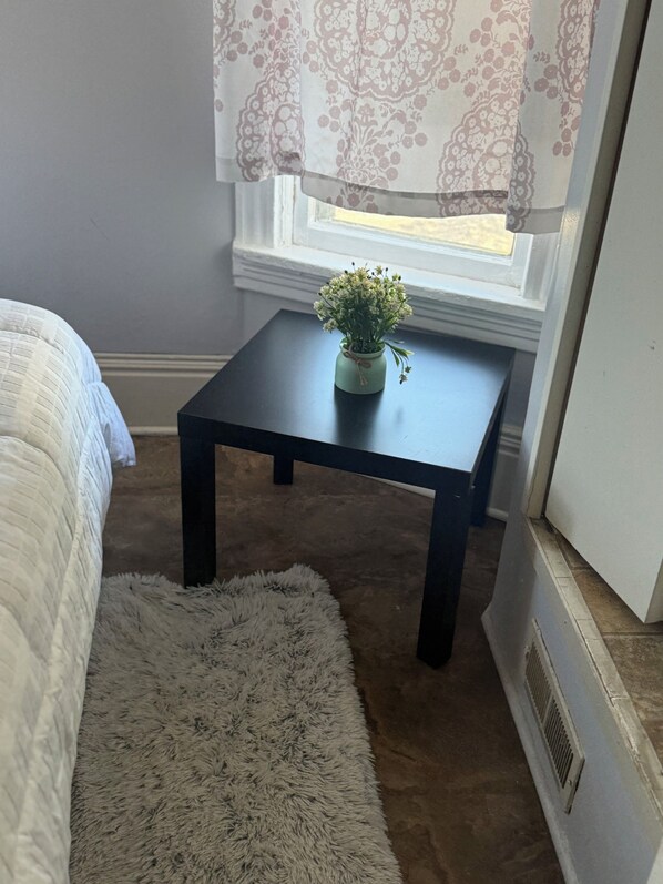 A bedroom with a bed, a small table with a plant, a fluffy rug, and a window with curtains.