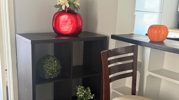 A corner of a room with a black shelving unit, a wooden chair, and a kitchen area with white cabinets.