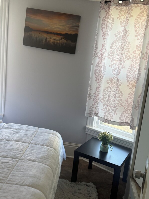 Une chambre à coucher avec un lit, une petite table sur laquelle est posée une plante, une fenêtre avec des rideaux à motifs et un tableau accroché au mur.