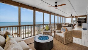 Un espace extérieur abrité avec vue sur la plage, une table ronde en verre bleu, des meubles en osier et un ventilateur de plafond.