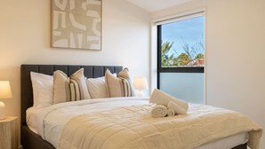A neatly made bed with a quilt, pillows, and rolled towels, a bedside lamp, and a window with a view of trees.