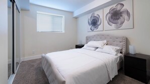 A bedroom with a bed, a nightstand, a window with blinds, and two wall-mounted flower paintings. - Heart of Corydon (Winnipeg)