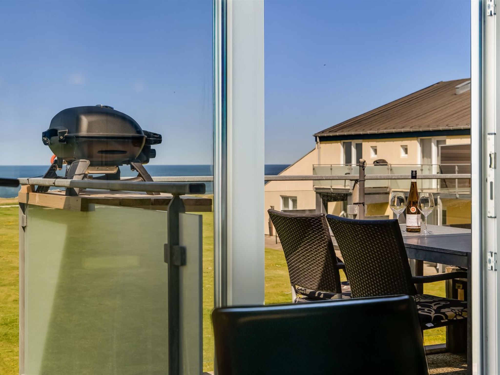 Vista de varanda com churrasqueira, cadeiras de vime e uma mesa com taças de vinho.
