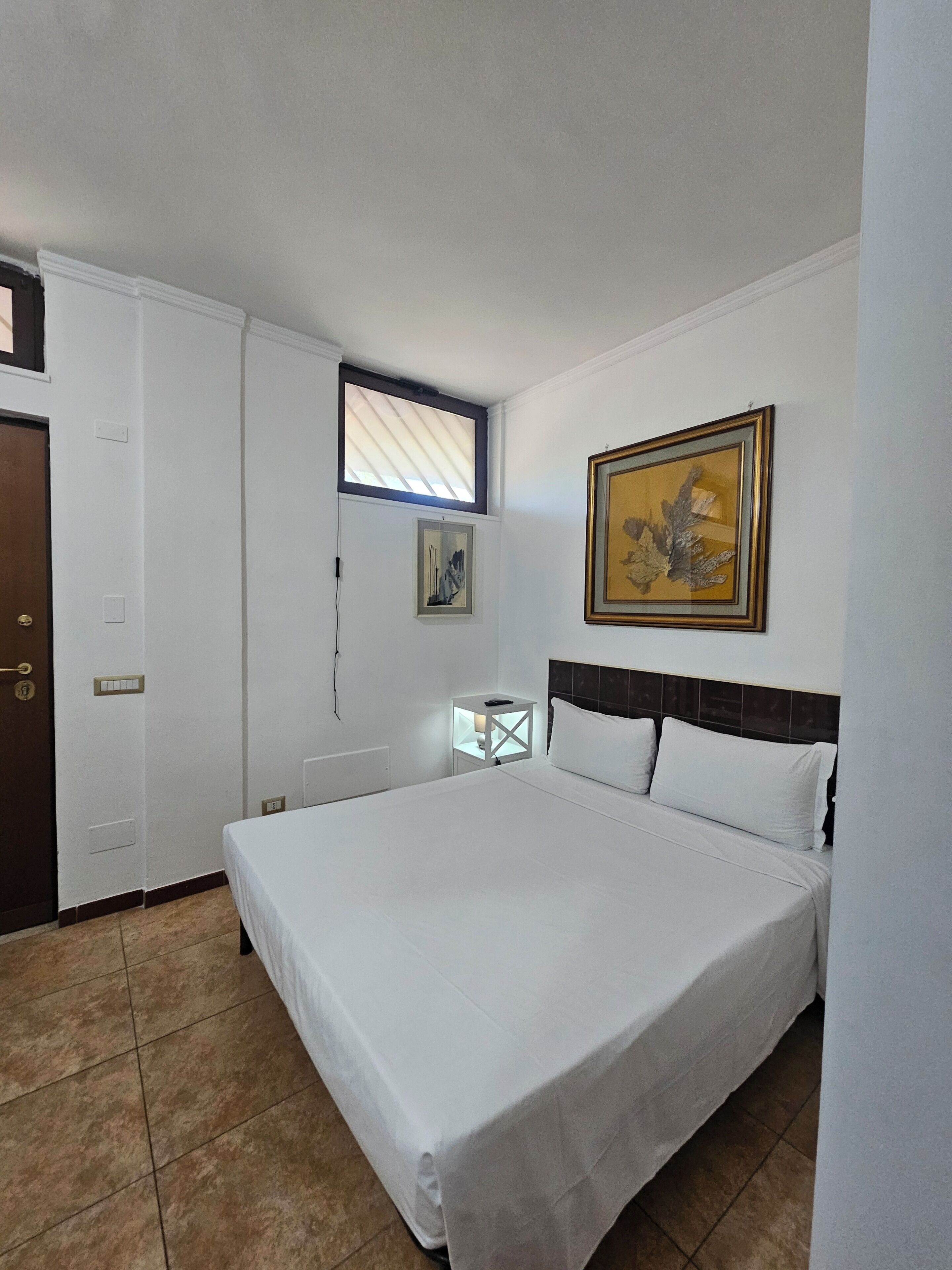 A bedroom with a bed, a nightstand, a window, and a painting on the wall.