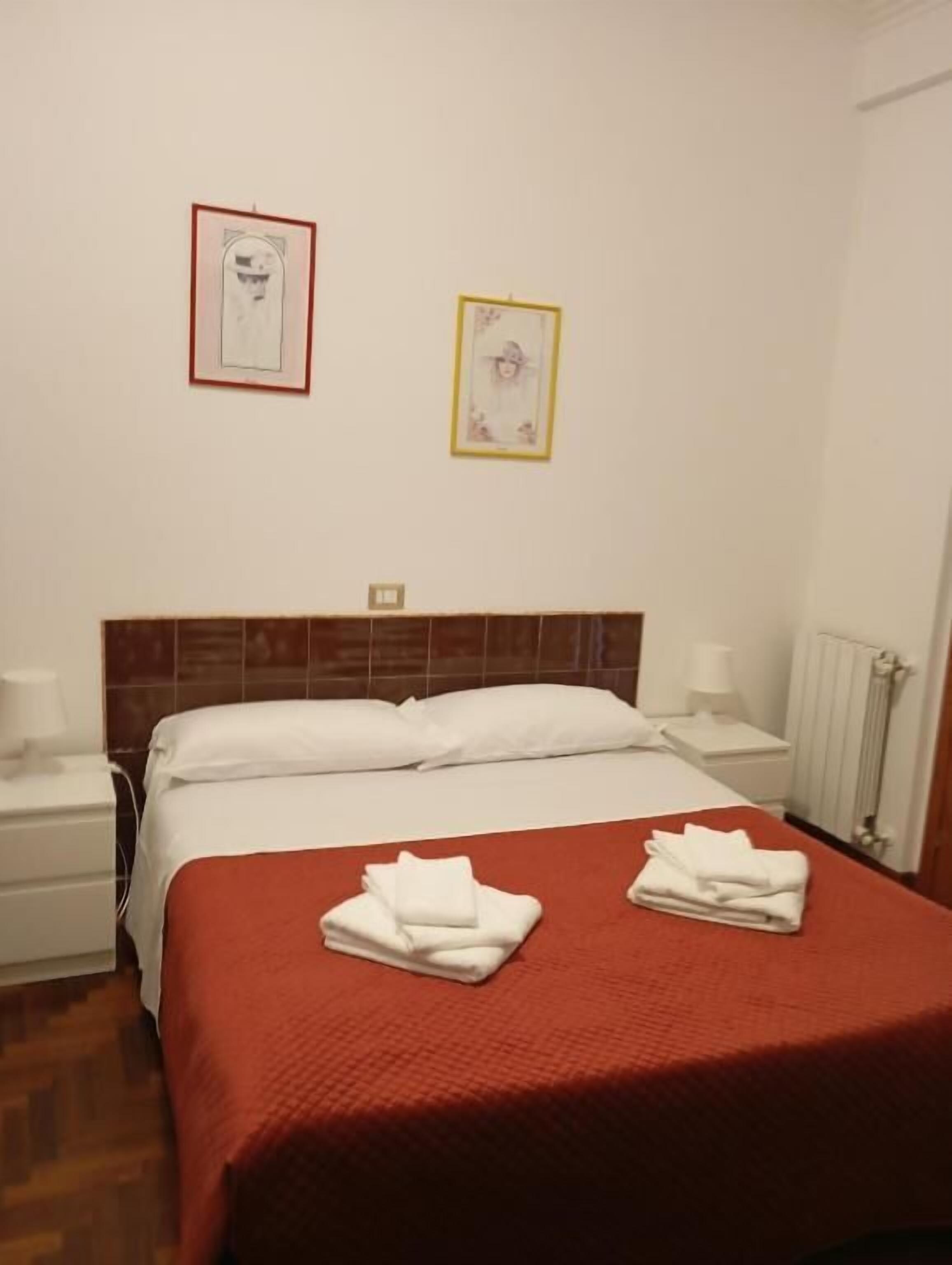 Una habitación de hotel con una cama, dos cuadros en la pared, una mesita de noche y un radiador.