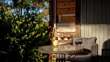 Mesa redonda com garrafa e duas taças de vinho, almofada e cadeira em área externa sombreada com paredes de madeira e vegetação.