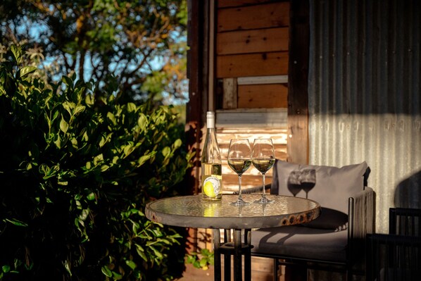 A round table with a bottle and two wine glasses, a cushion, and a chair in a shaded outdoor area with wooden walls and greenery.