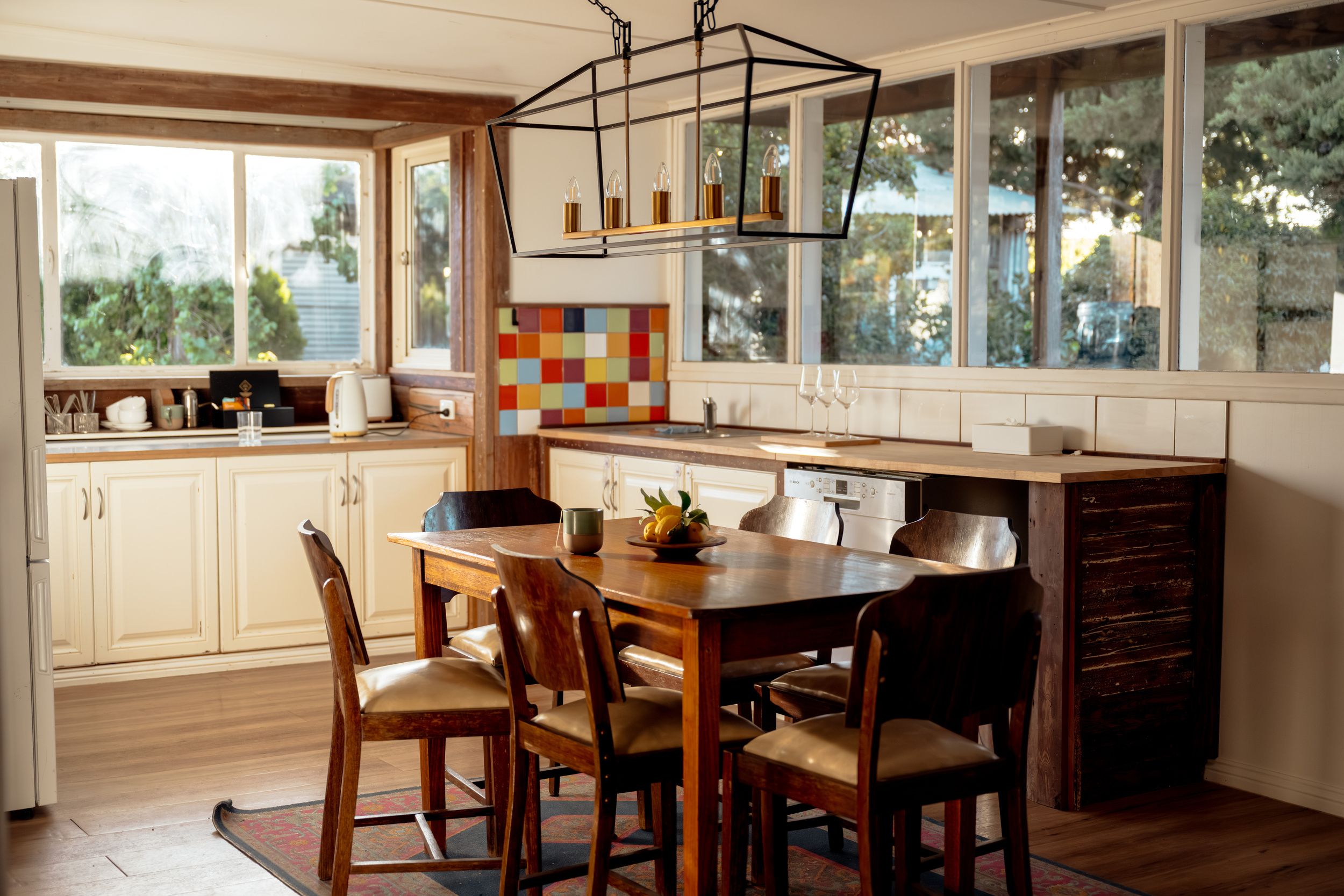 A kitchen with wooden cabinets, a dining table with chairs, a colorful backsplash, and a large window with a view of trees.