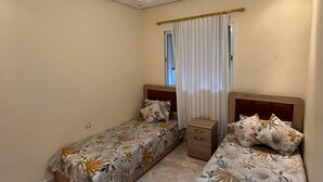 A small, well-lit room with two single beds, a wooden nightstand, and a window with curtains. - Close to the beach—3-minute drive—Secure apartment complex with a pool (Bouznika)