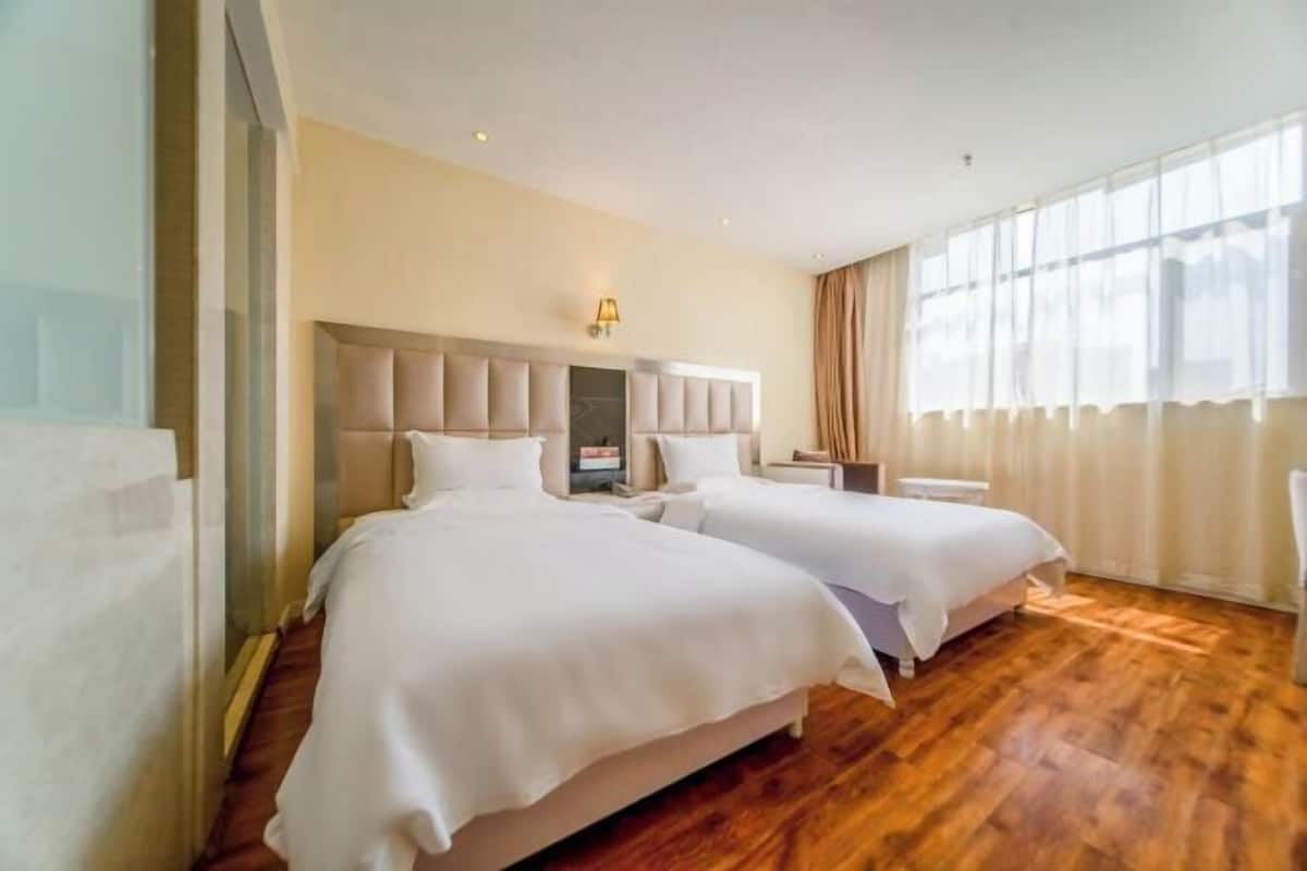 A hotel room with two beds, a wooden floor, a headboard with a light fixture, and a large window with curtains.