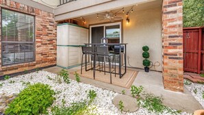A patio with a table and chairs, a ceiling fan, string lights, and a small garden area with plants and pebbles. - Modern 2BR Condo, Fast Wi-Fi, Blackout Draperies (Irving)