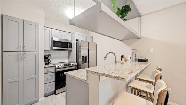 A modern kitchen with grey cabinets, a stainless steel refrigerator, a built-in microwave, and a granite countertop. - Modern 2BR Condo, Fast Wi-Fi, Blackout Draperies (Irving)