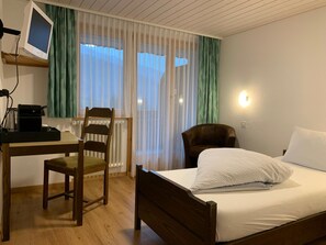 A hotel room with a wooden desk, a chair, a bed with white bedding, a television on a stand, and a window with sheer curtains.