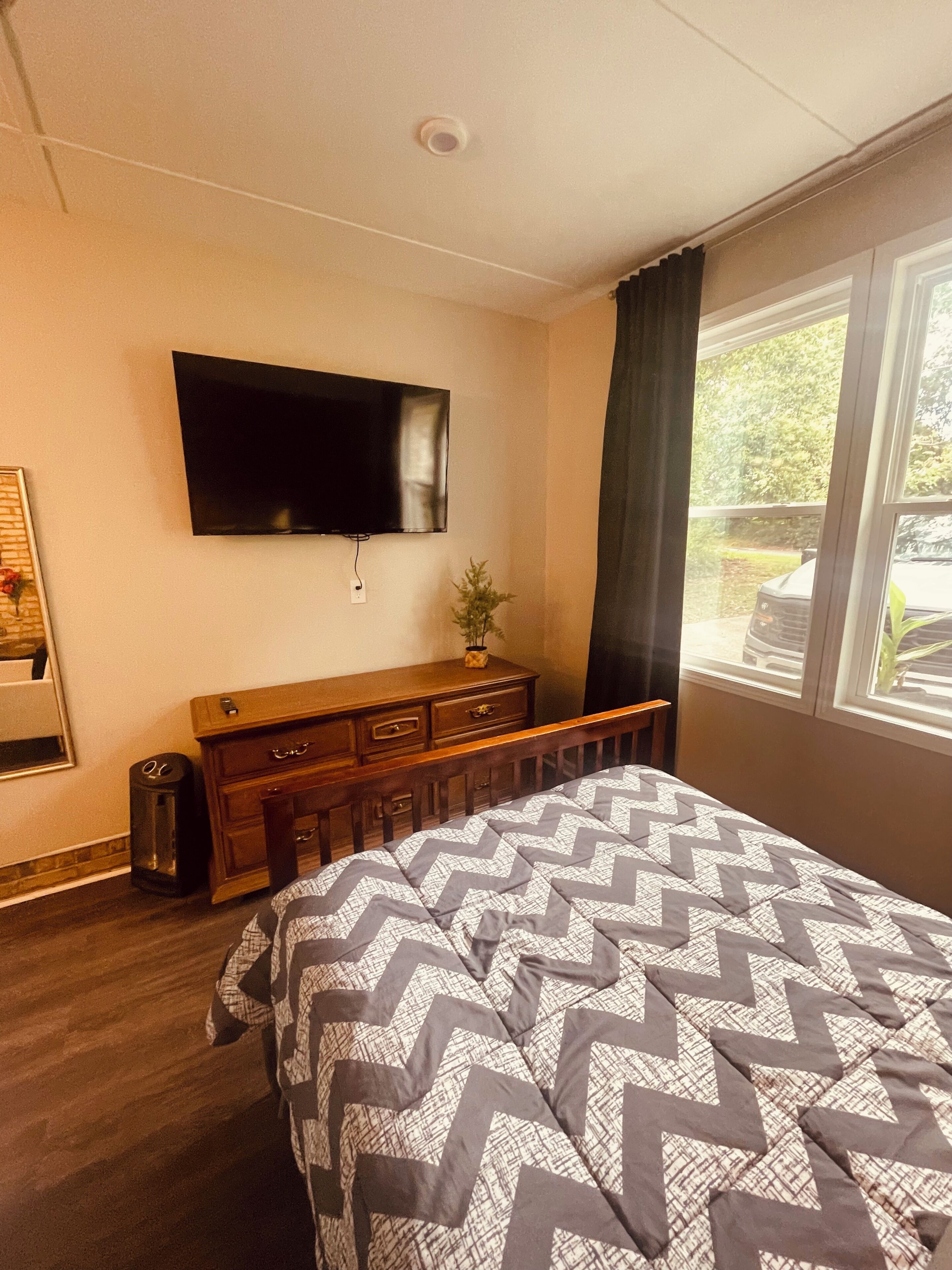 A bedroom with a bed, a TV mounted on the wall, a dresser, and a window with a view of greenery.