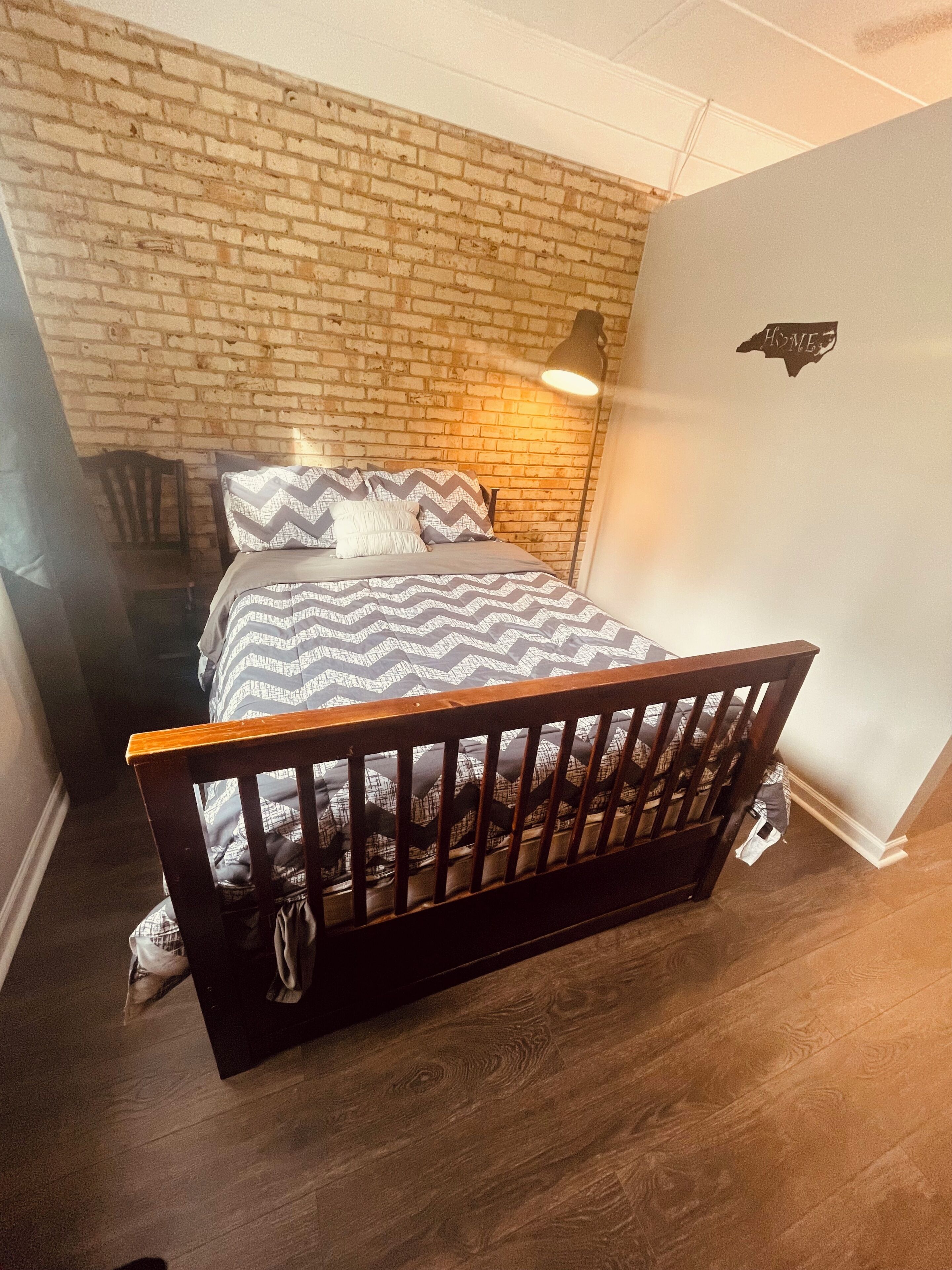 Een slaapkamer met een bakstenen muur, een bed met houten hoofdeinde, een vloerlamp en een bordje aan de muur.