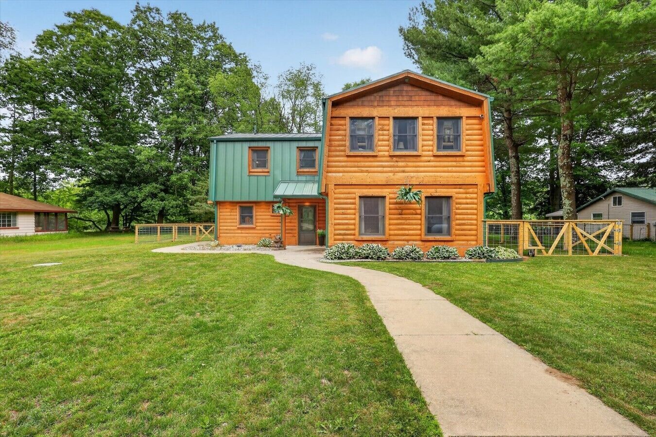 A two-story log cabin with a green roof, a concrete pathway leading to the entrance, and a fenced yard.