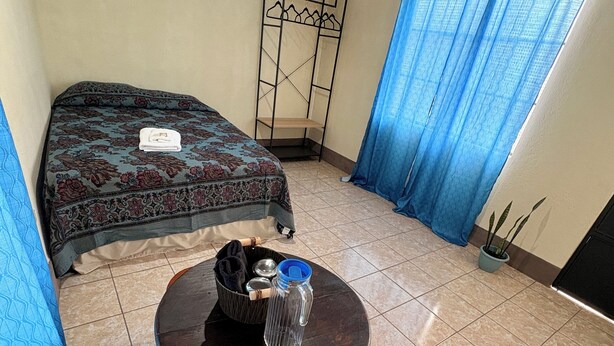 A room with a bed, a small table with a water bottle, and a blue curtain. - Casa de Huéspedes Elizabeth (Antigua Guatemala)