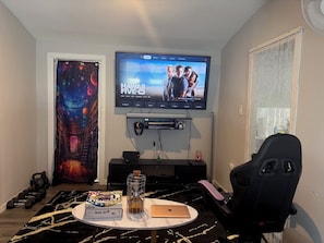 Una sala de estar con una televisión de pantalla plana montada en la pared, una silla gamer, una mesa de centro con una computadora portátil y una estantería.