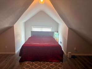 Un dormitorio con techo inclinado, cama con colcha roja, ventana con persianas y un cartel en la pared.
