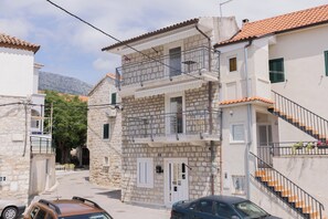 Ein zweistöckiges Steinhaus mit Balkon und Treppe, vor der ein Auto geparkt ist.