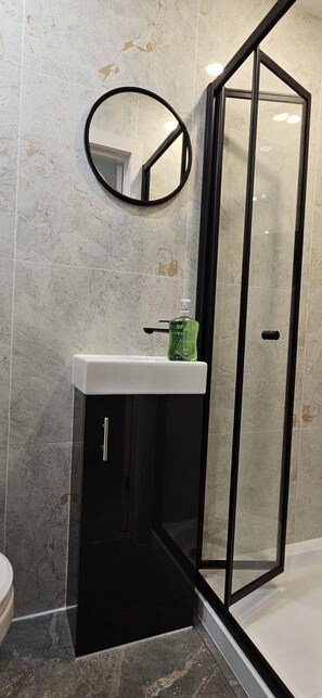 Ein Badezimmer mit Duschkabine aus Glas, einem runden Spiegel und einem weißen Waschbecken mit einer grünen Flasche auf der Ablagefl äche.